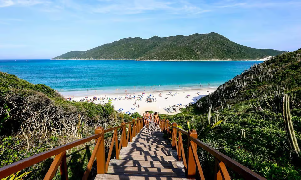 Wooden staircase leading to a beautiful turquoise beach with white sand, surrounded by green hills and cacti