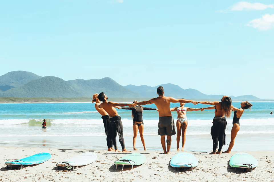 Tourism in Brazil: Group of surfers practicing stretches on a scenic Brazilian beach with surfboards, mountains, and blue ocean in the background