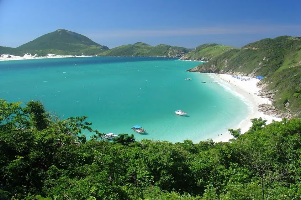 Tourism in Brazil A breathtaking view of a turquoise beach surrounded by lush green hills, with boats floating on the clear water and white sand in the distance